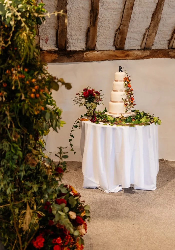 A four-tier white wedding cake decorated with orange and pink flowers, placed on a white-cloth-covered table with green foliage, in a rustic setting with wooden beams and a textured white wall.