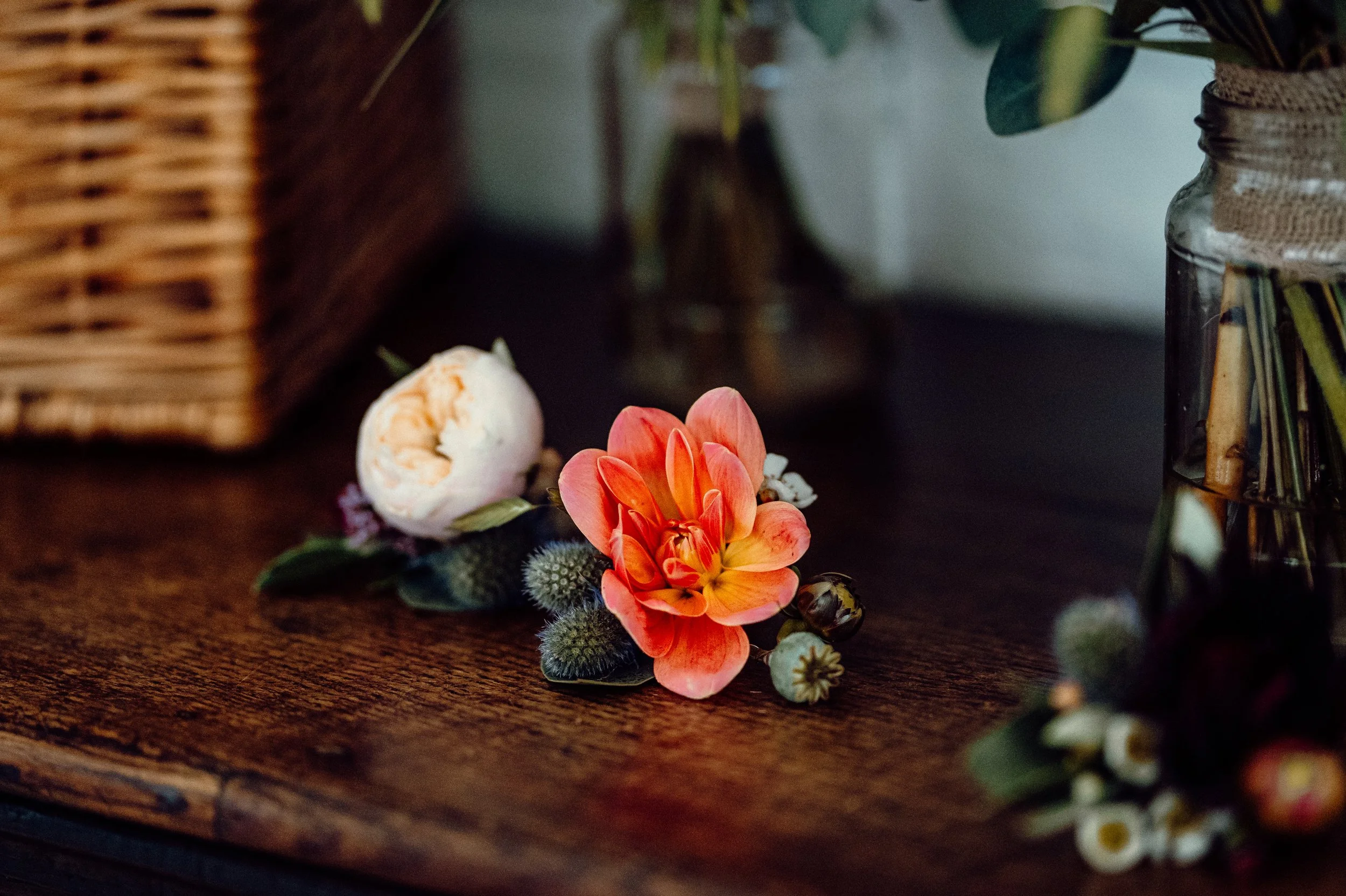 A peony flower, some berries, and a closed peony bud arranged on a dark wooden surface with a glass vase and wicker basket in the background.
