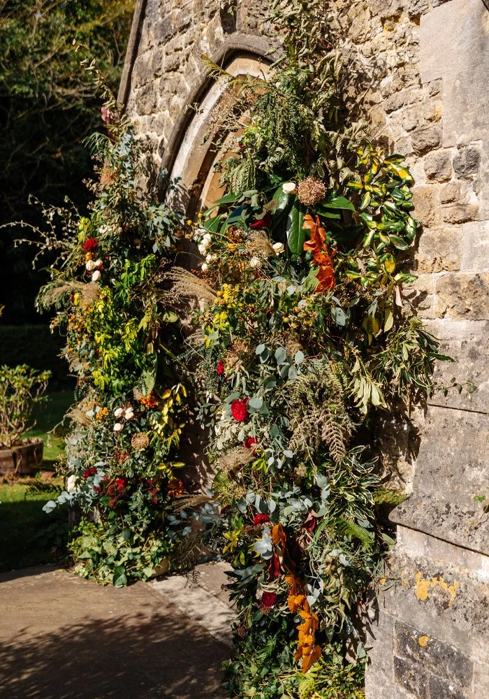 A floral wreath made of various green leaves and colorful flowers hanging on a stone building exterior.