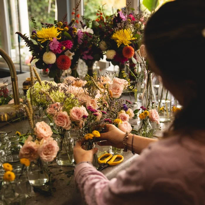 Person arranging pink roses and yellow flowers in small glass vases, with more floral arrangements on the table