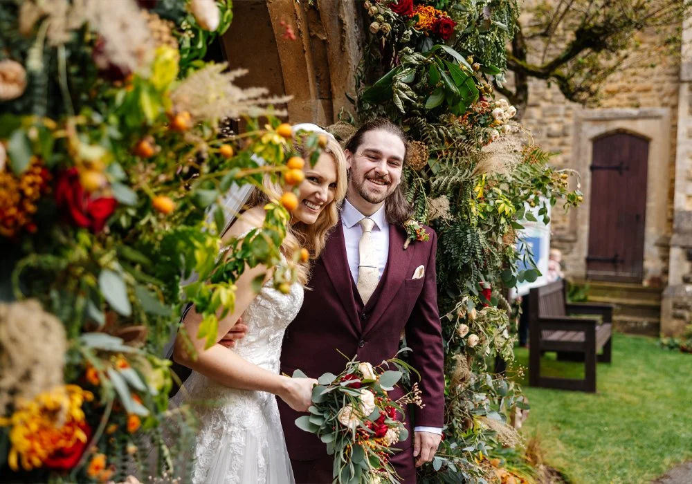 A bride and groom smiling and standing together outdoors, framed by a lush floral arrangement, with a stone building and greenery in the background.