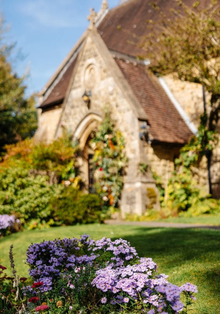 A stone church with a steep roof and arched doorway surrounded by colorful flowers and green foliage on a sunny day.