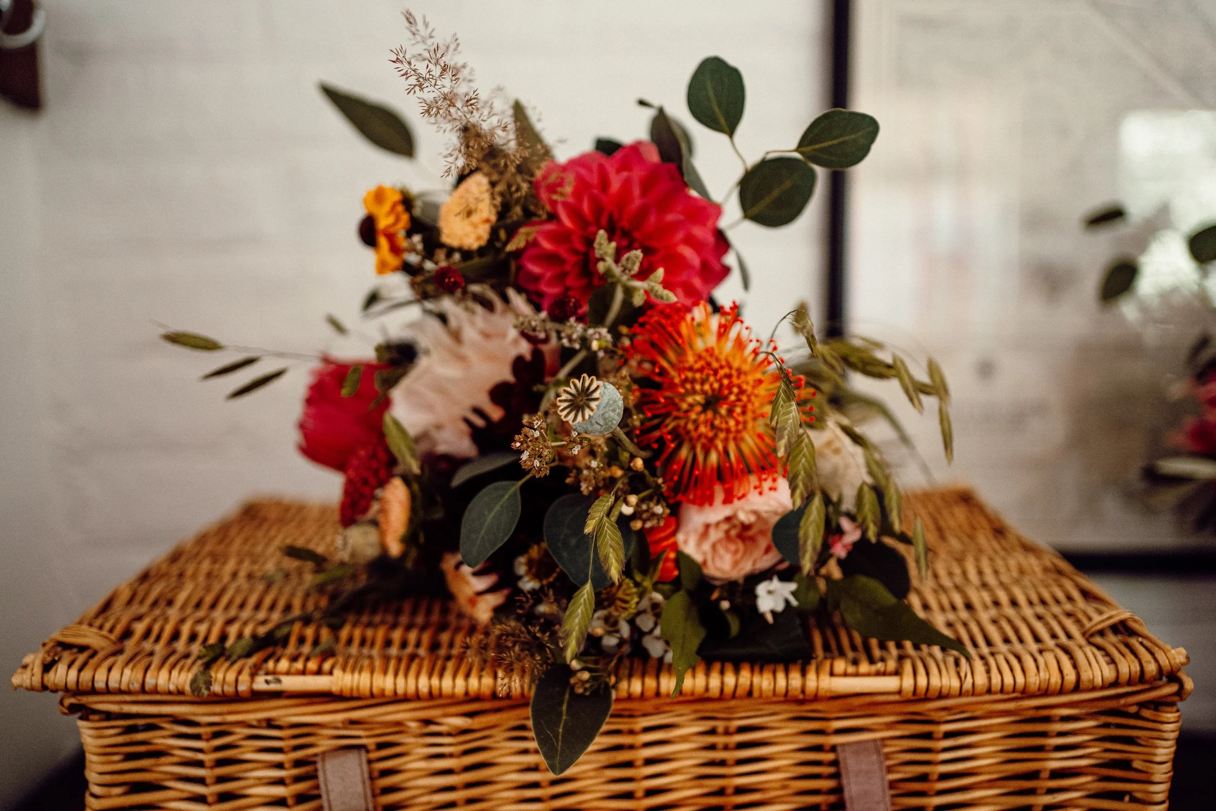 A colorful bouquet of flowers, including pink, red, orange, and white blooms, with green foliage, placed on a woven wicker surface.