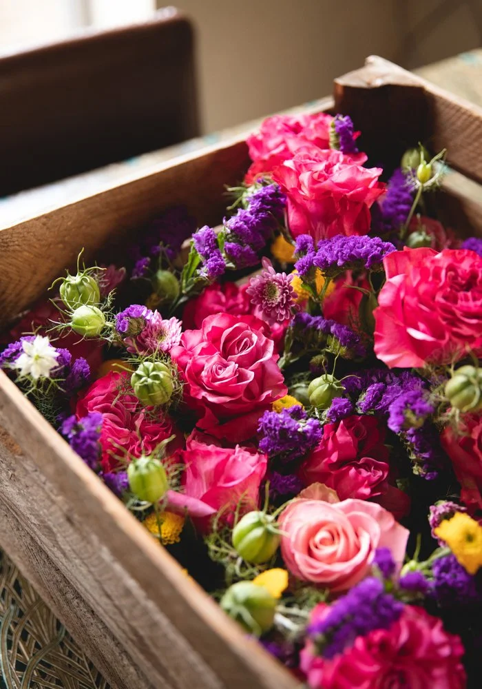 A wooden crate filled with pink roses, purple statice, green buds, and small yellow and white flowers.