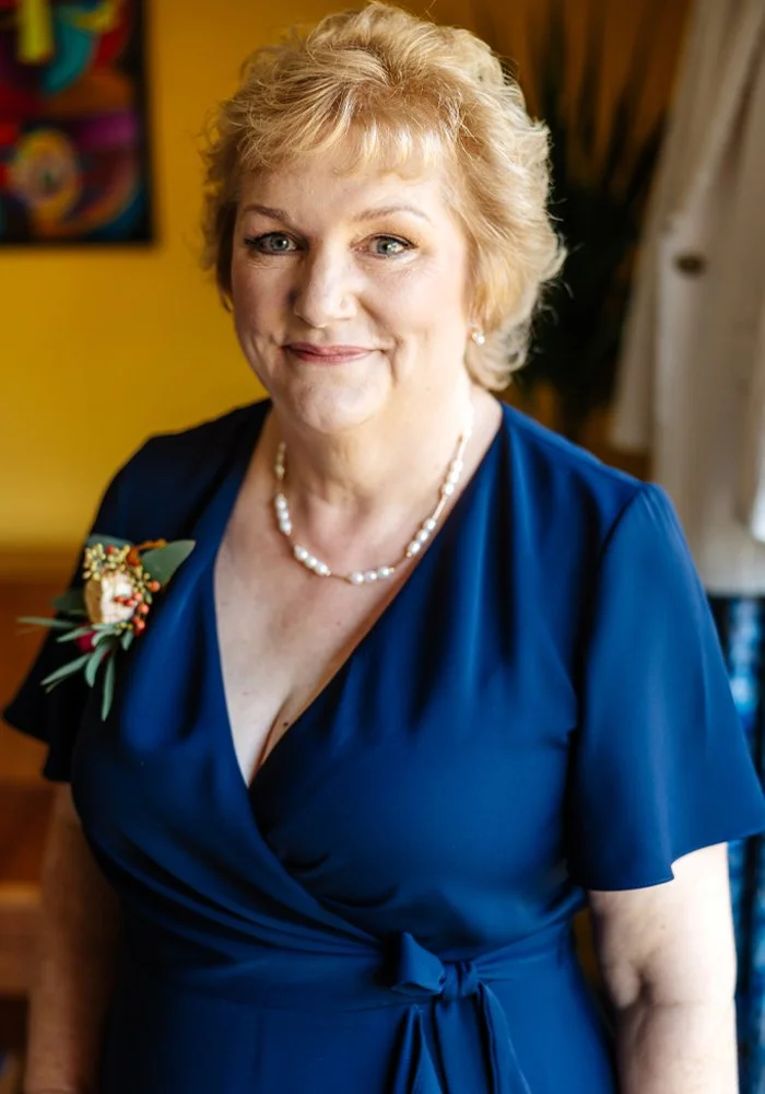 A smiling woman with blonde hair in a blue dress, standing indoors with a yellow wall and art in the background, wearing a pearl necklace and a corsage on her left shoulder.