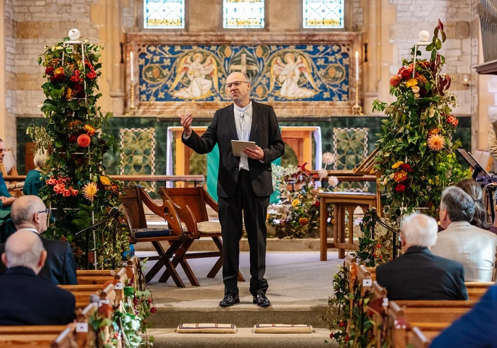 A man in a black suit giving a speech in a church decorated with flowers and greenery.