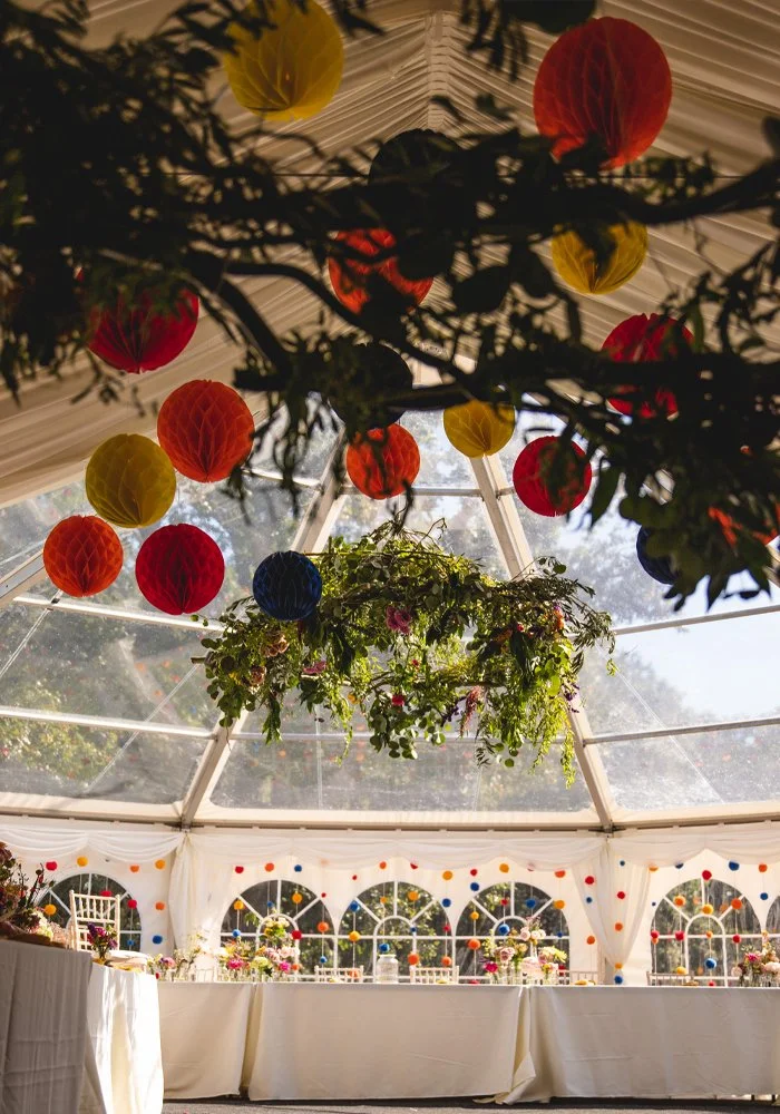 Decorative hanging paper lanterns in red, yellow, blue, and orange hues surround a floral centerpiece beneath a clear-paneled tent ceiling with draped white curtains and string lights, set up for an outdoor celebration or event.