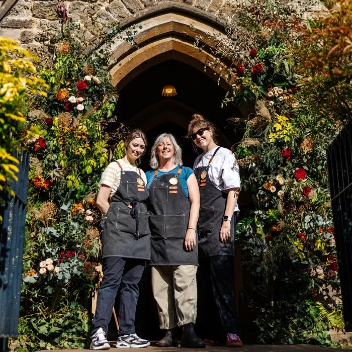 Three women standing together under a floral archway outside, smiling, wearing black aprons, with one woman in the middle and two women on either side.
