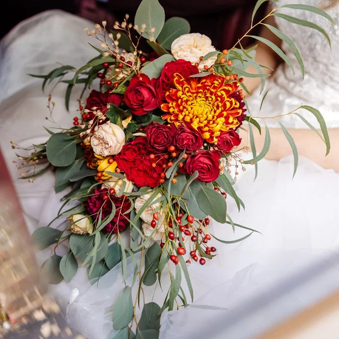 A bridal bouquet with red roses, a yellow and red chrysanthemum, white flowers, and assorted greenery and berries.