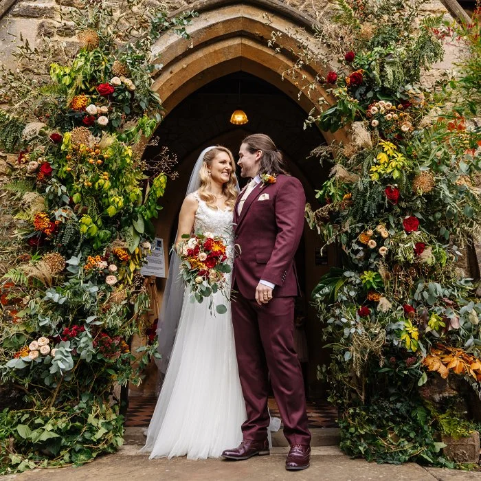 A bride and groom stand together under a stone archway decorated with colorful flowers, smiling at each other after their wedding ceremony.