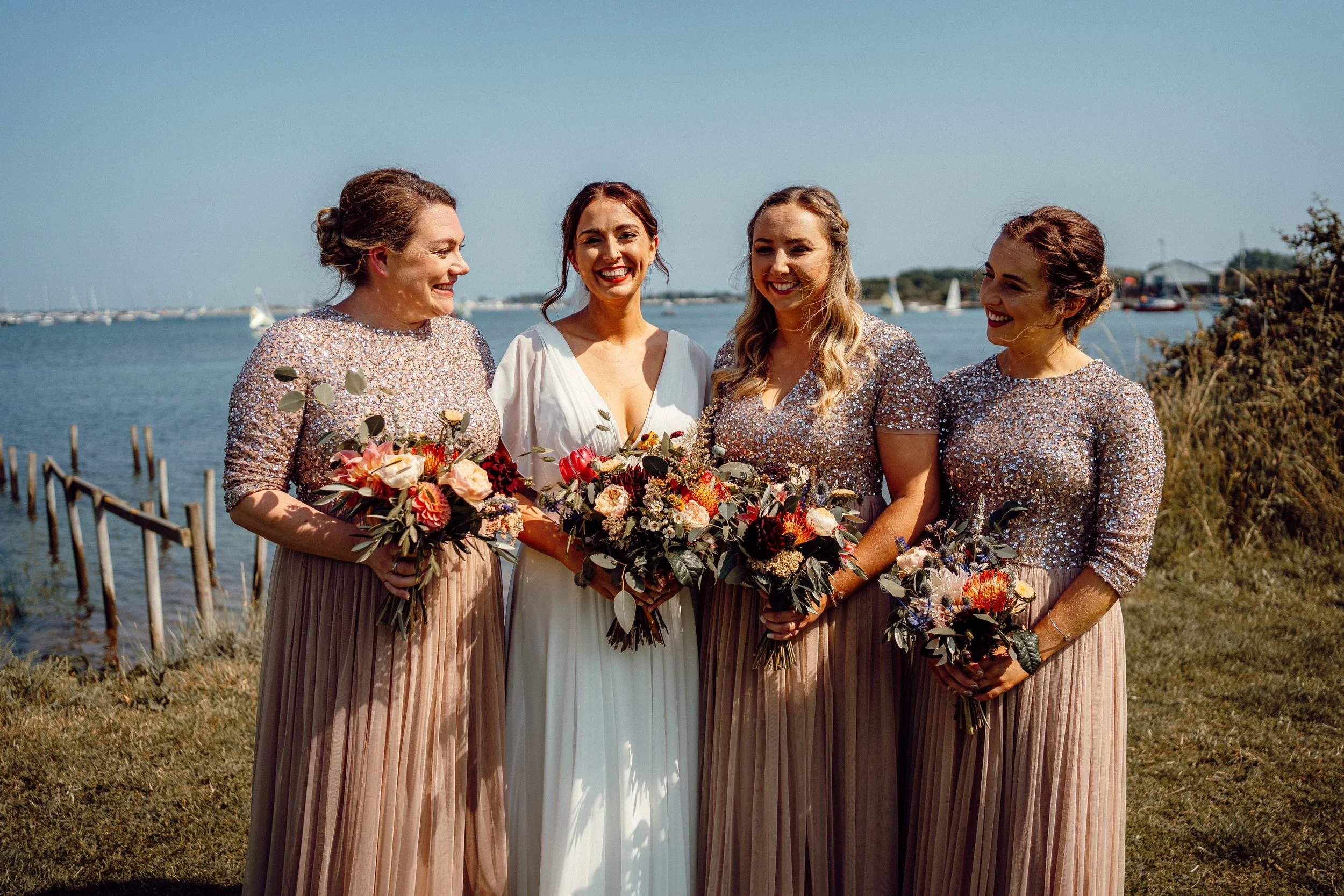 Four women in formal dresses holding bouquets, standing outdoors by water, smiling and enjoying a sunny day.