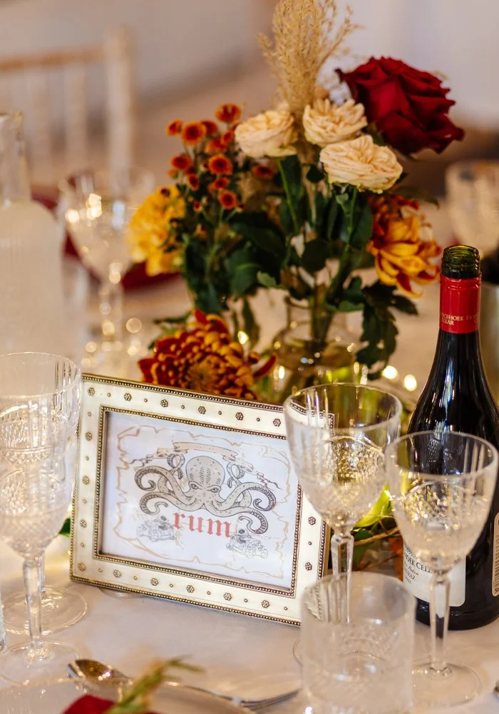 Elegant table setting with a floral centerpiece, glassware, and a framed place card labeled 'rum' at a formal event or celebration.