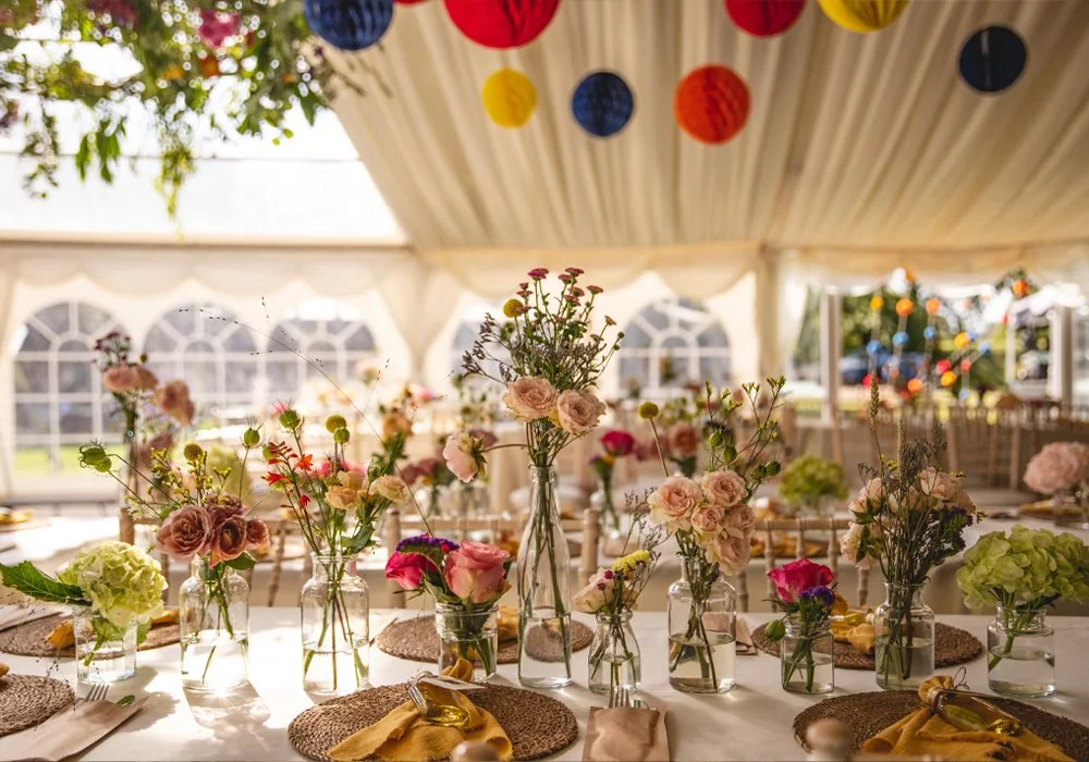 Decorated event tent with floral centerpieces in glass vases and colorful hanging paper lanterns.