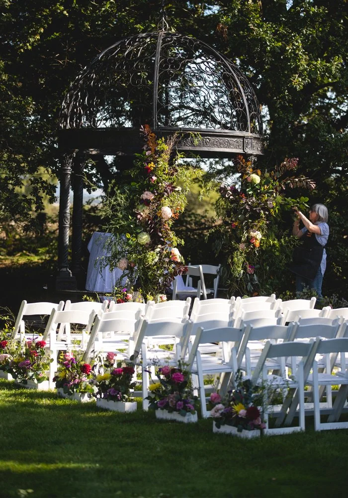 Outdoor wedding ceremony setup with white chairs and floral arrangements, a decorative metal pavilion, and a person adjusting flowers.