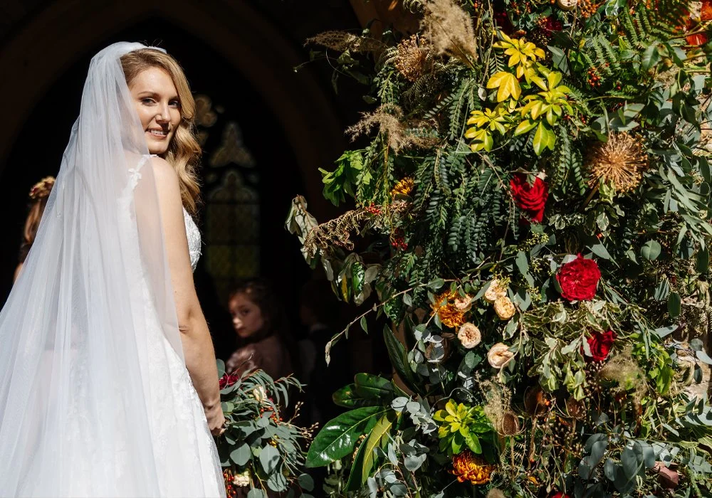 A bride in a wedding dress and veil smiling at the camera at her wedding ceremony, standing next to a large floral arrangement with various flowers and greenery.