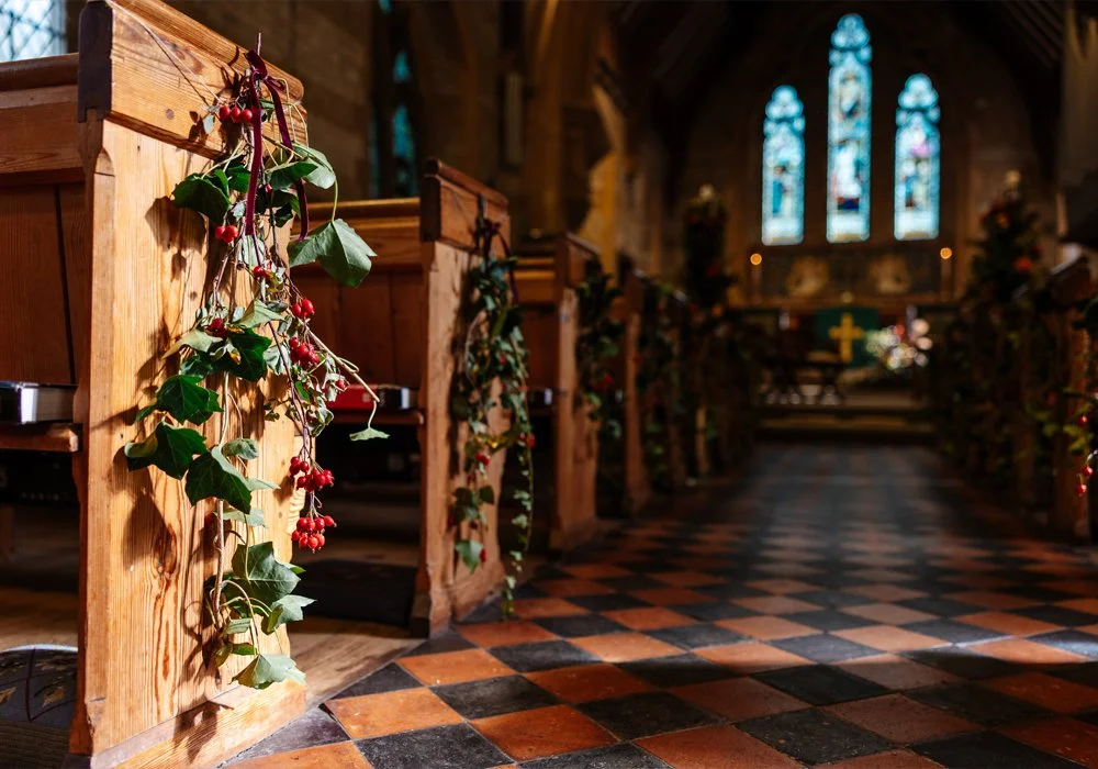 Interior of a church decorated with green ivy and red berries on the pews, with stained glass windows and an altar in the background.