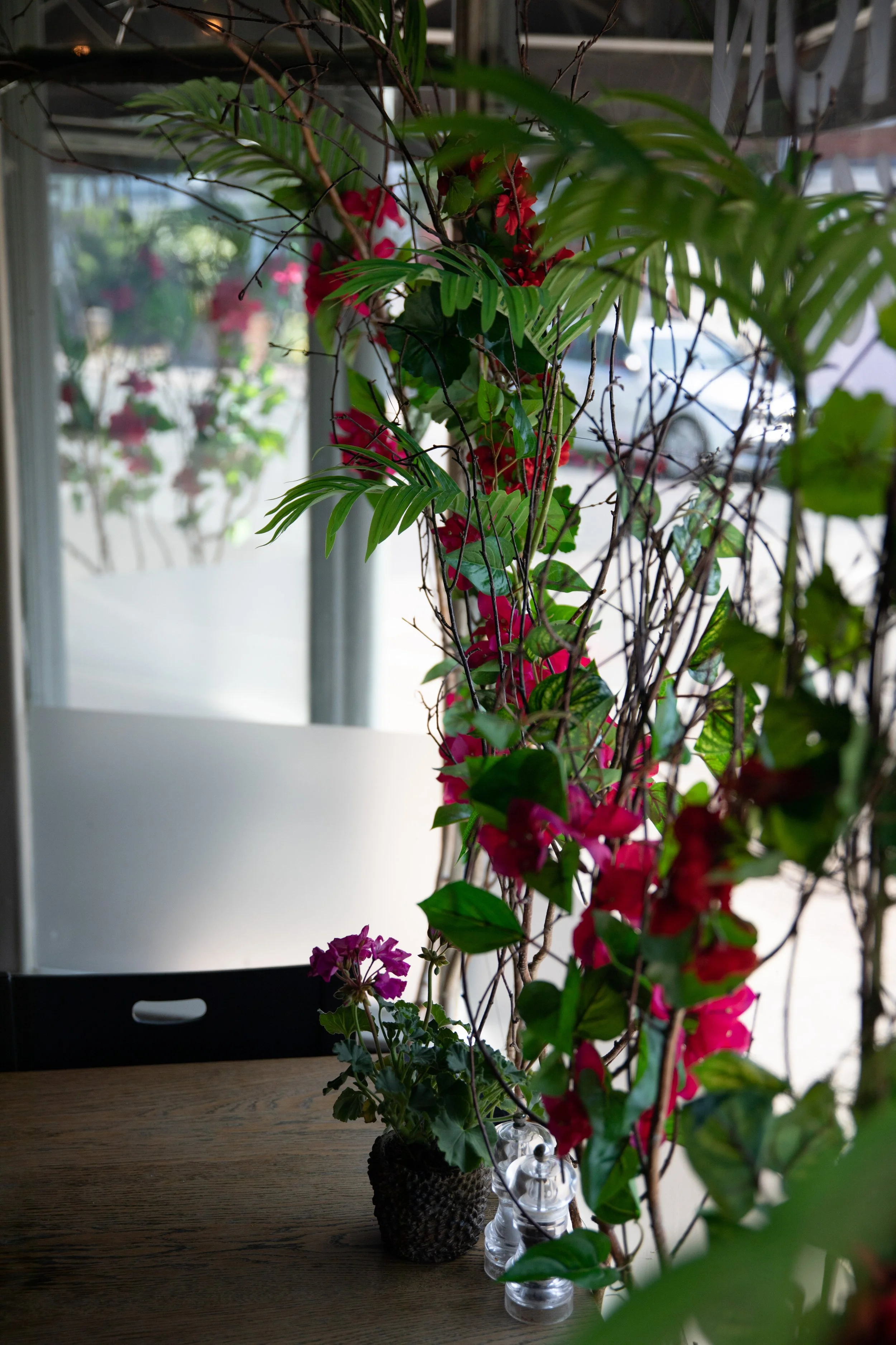 Indoor scene with vibrant pink and purple flowers, green leaves, and branches near a window, with a wooden table and a black chair.