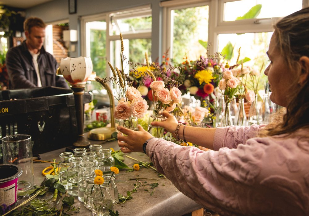 A woman arranging pink and peach flowers on a table filled with floral supplies, with a man working at a piano in the background inside a bright room.