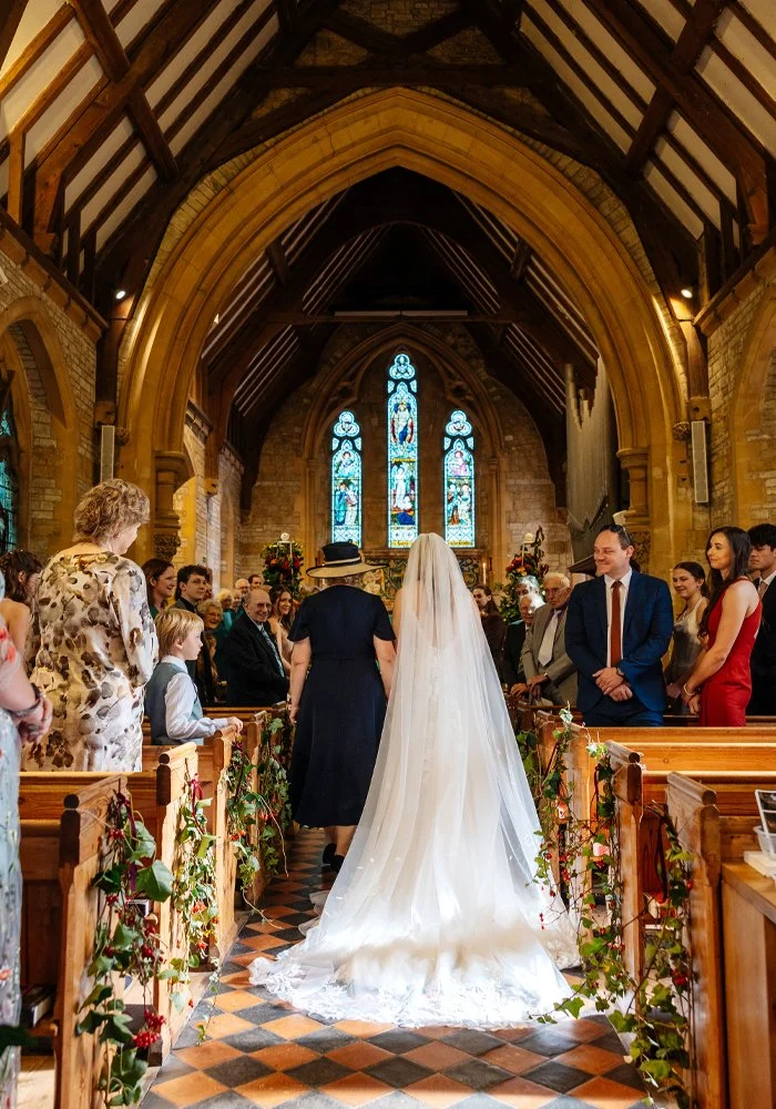 A bride in a white wedding dress with a long train and veil walking down the aisle of a church, flanked by guests on either side, with stained glass windows and wooden architecture in the background.