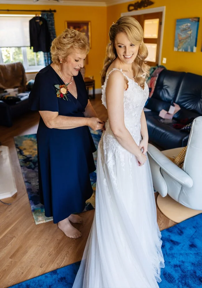 A young woman in a white wedding dress smiling as an older woman helps her with her dress in a warmly decorated living room.