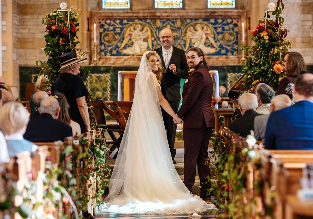 A bride and groom holding hands at their wedding ceremony inside a church, with an officiant and guests in attendance.