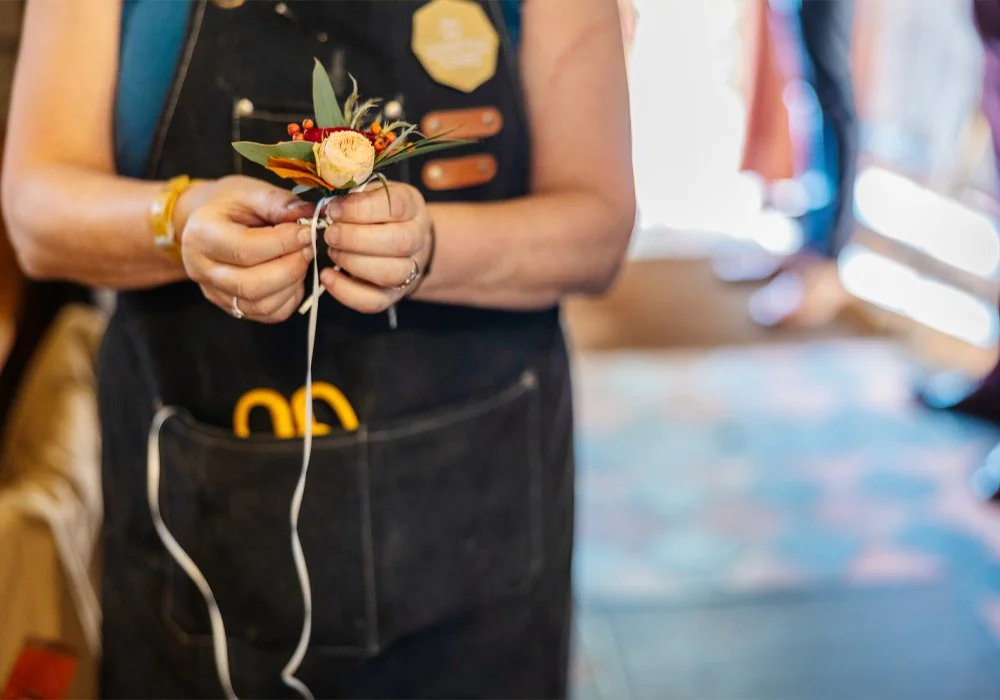 A person holding a small floral boutonniere, wearing a black apron, with a yellow bracelet and scissors in the apron pocket, inside a brightly lit room.