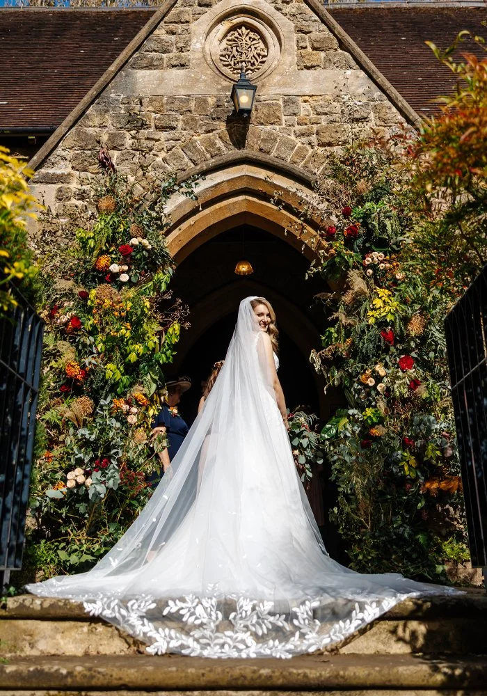 Bride in a wedding dress and veil standing at a Hampshire church entrance, framed by Autumnal flower installation..