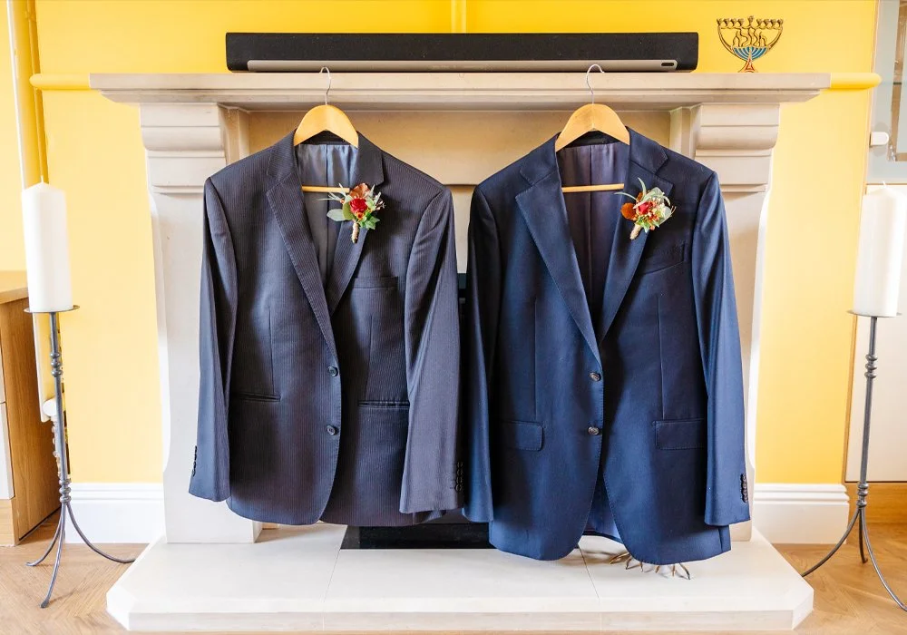 Two suits with boutonnieres hanging on hangers in front of a fireplace.