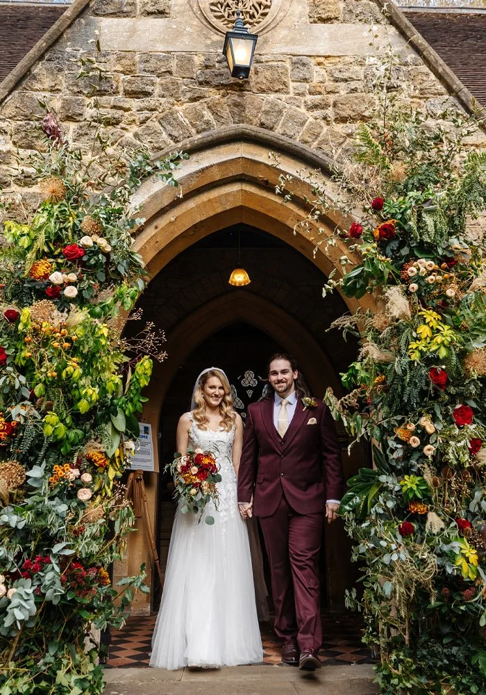 Bride and groom walking out of a church decorated with colorful flowers and greenery, holding hands, smiling, during their wedding.