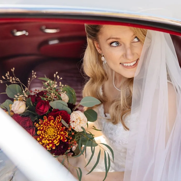 Bride with a bouquet of red, pink, and yellow flowers, wearing a wedding dress and veil, sitting inside a car, smiling and looking out.
