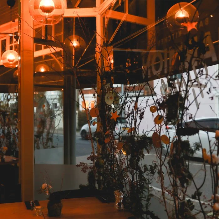 Interior of a cafe with orange lighting, hanging light fixtures, and decorative branches with star and circle ornaments near the window, with parked cars visible outside.