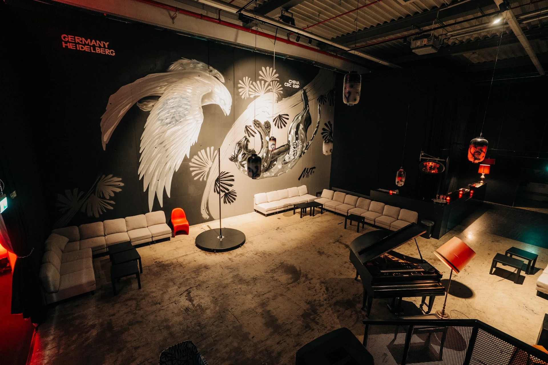 Interior of a lounge or club with beige sectional sofas, a black grand piano, lamps, and a large mural of an eagle wing, floral patterns, and a colorful depiction of a dragon dance, with words 'Germany Heidelberg' and 'China CDC GDP' on the mural.
