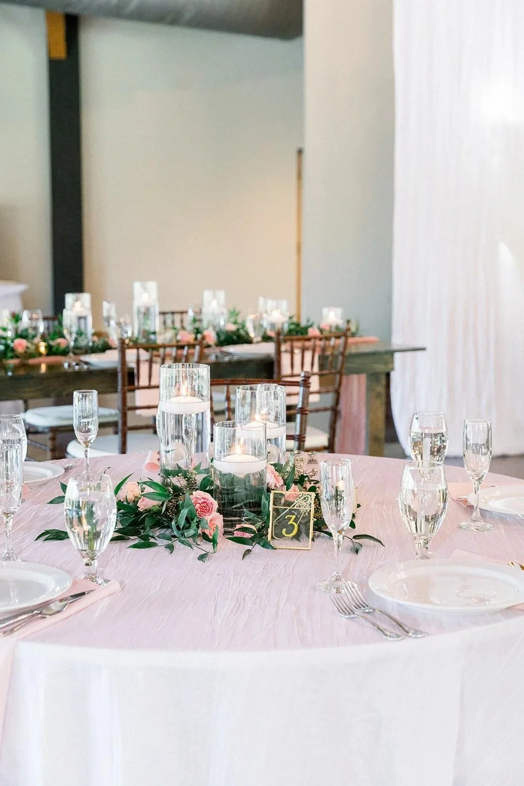 Elegant wedding reception table decorated with pink flowers, greenery, tall candles in glass holders, and glassware, with a floral centerpiece and a table number 3 card.