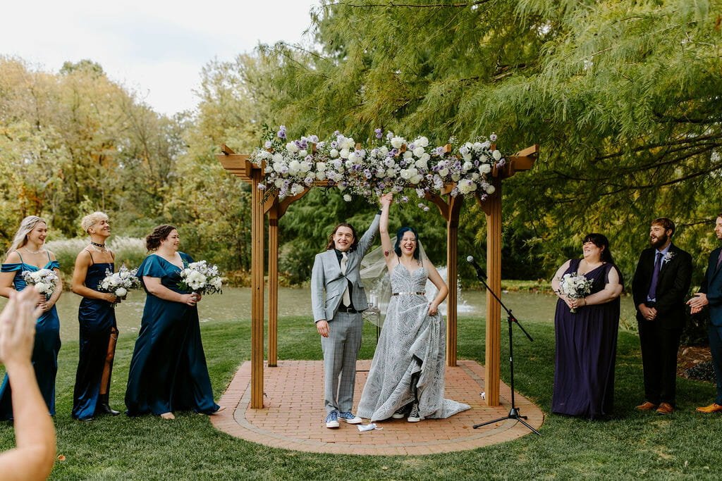 A joyful wedding scene outdoors with a couple under a floral wooden arch, surrounded by their smiling wedding party. The bride raises her arm triumphantly.