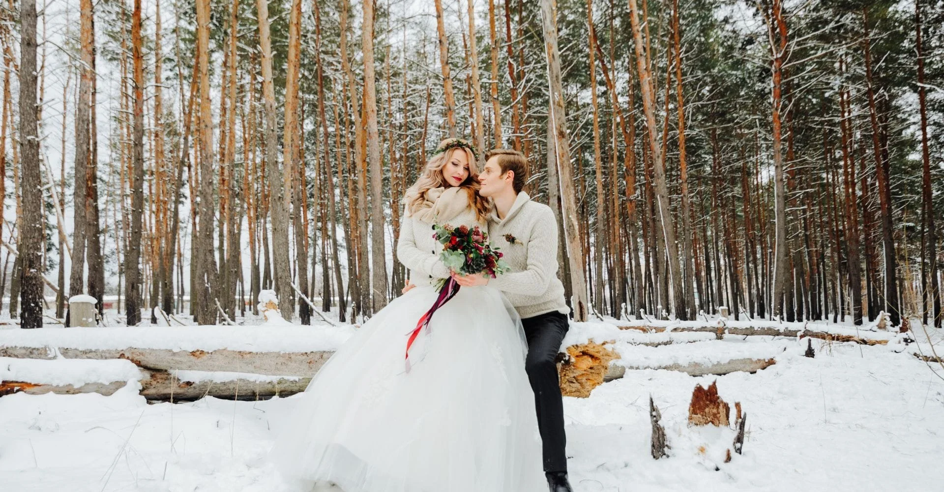 A couple embraces in a snowy forest. The woman wears a white wedding dress and holds a bouquet, while the man wears a light sweater, both smiling joyfully.