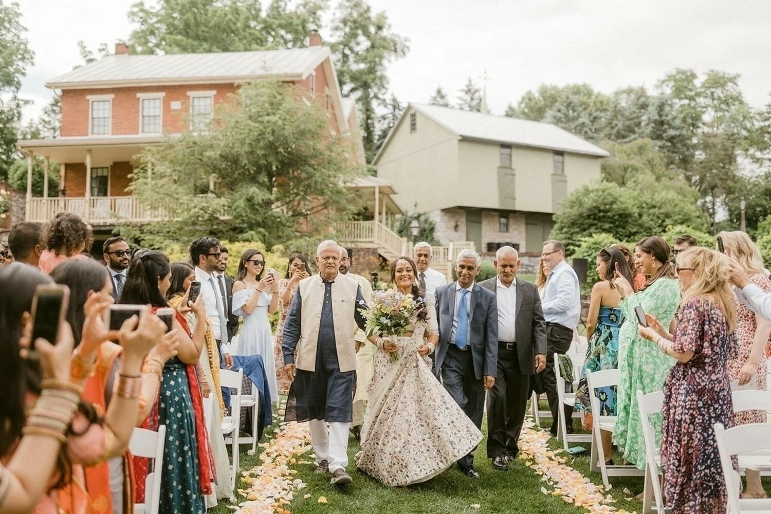 Bride in a floral dress walks down a flower-petal aisle, flanked by three older men. Guests in colorful attire are seated outdoors, capturing the moment.
