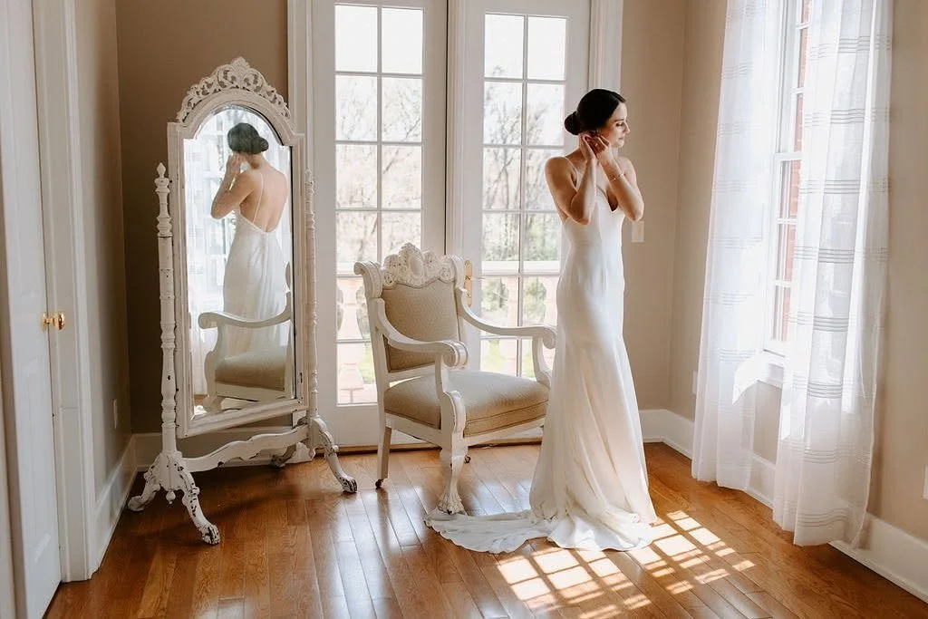 A bride in a stunning white dress admires her reflection in a mirror, capturing a moment of joy and anticipation.