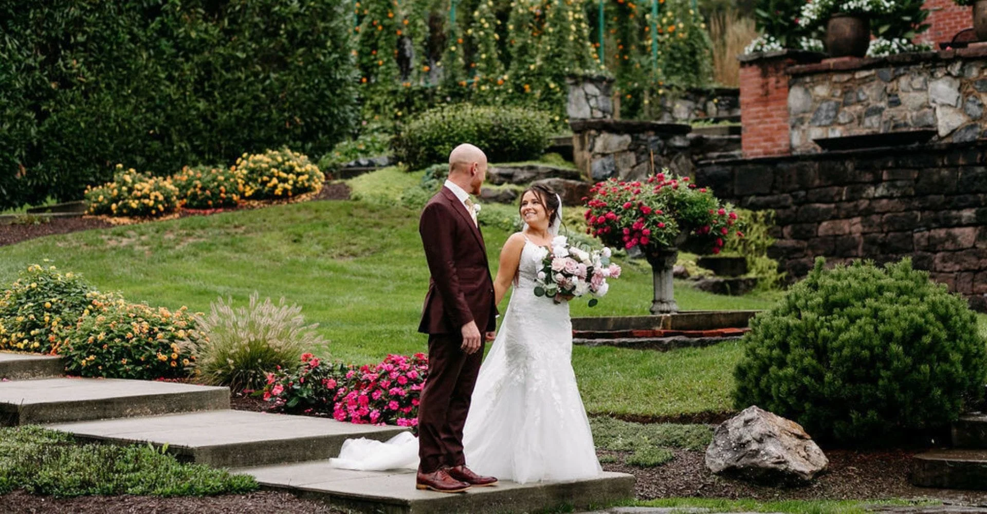 Bride in white gown holding bouquet and groom in dark suit standing on garden pathway surrounded by colorful flowers and greenery.