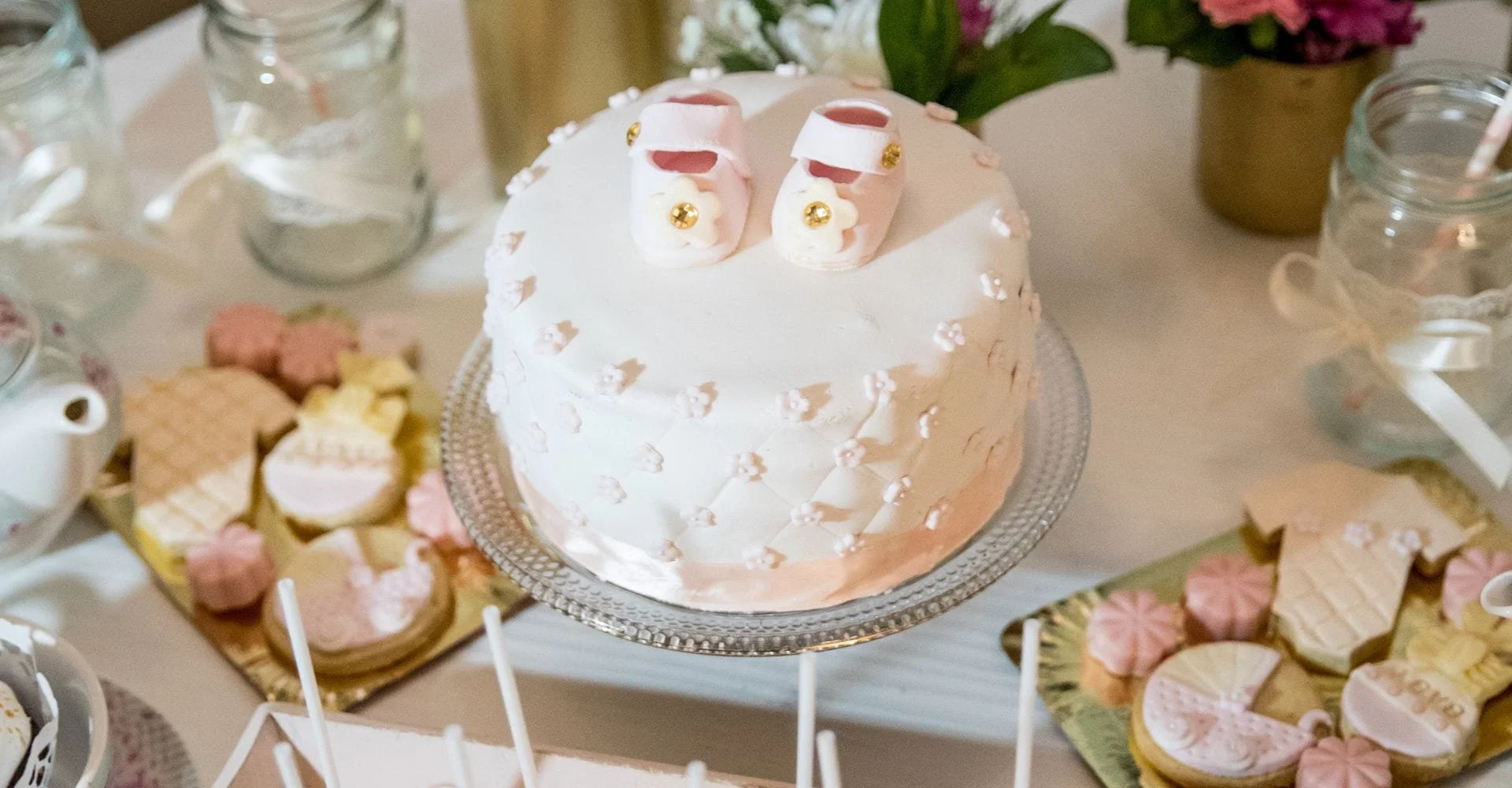 A white baby shower cake with tiny pink booties on top is the centerpiece. Surrounding it are pastel cookies and decorations, creating a festive atmosphere.