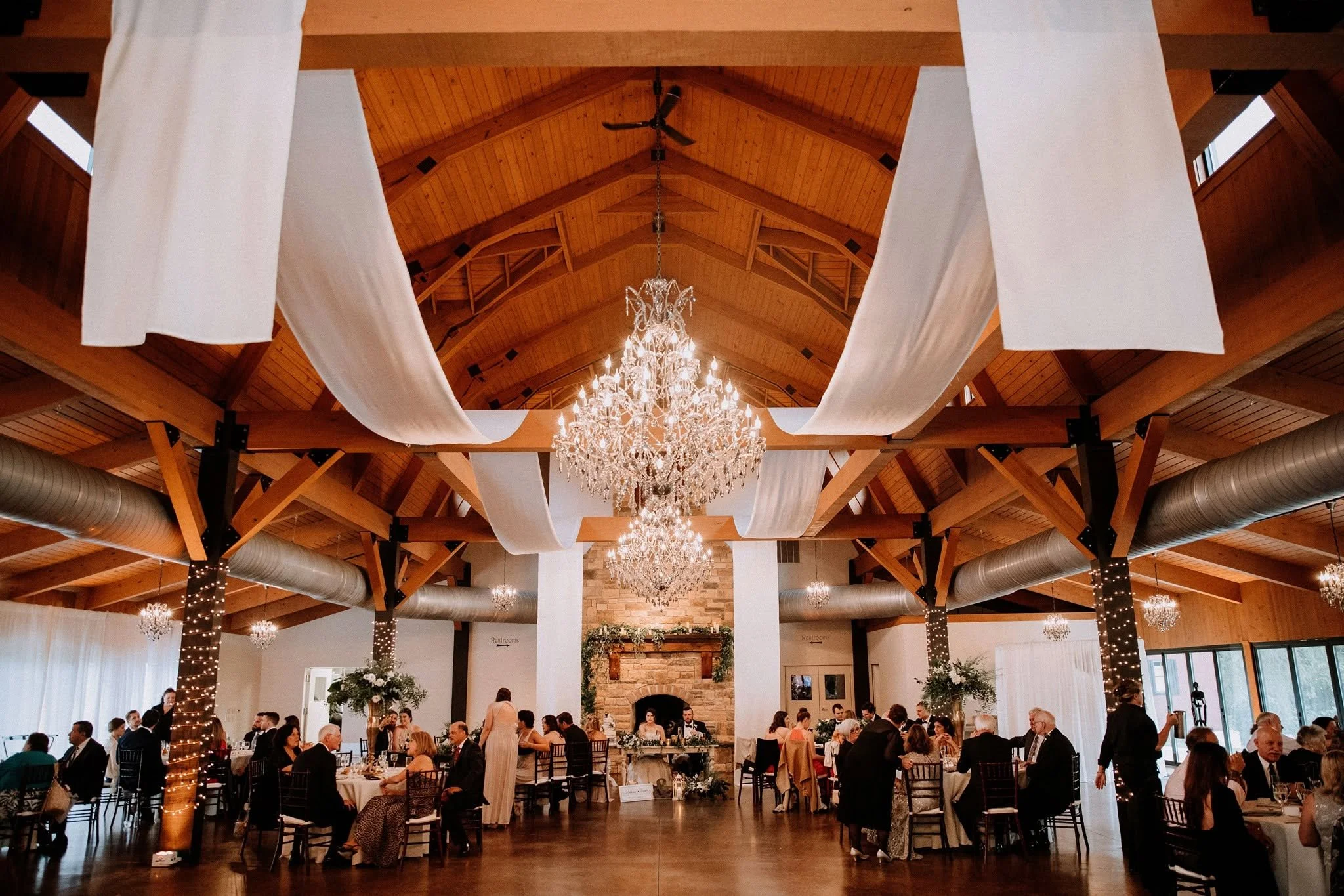 A warmly lit wedding reception fills a timber-vaulted hall, with flowing white drapes, glowing chandeliers, elegant tables and gathered guests.