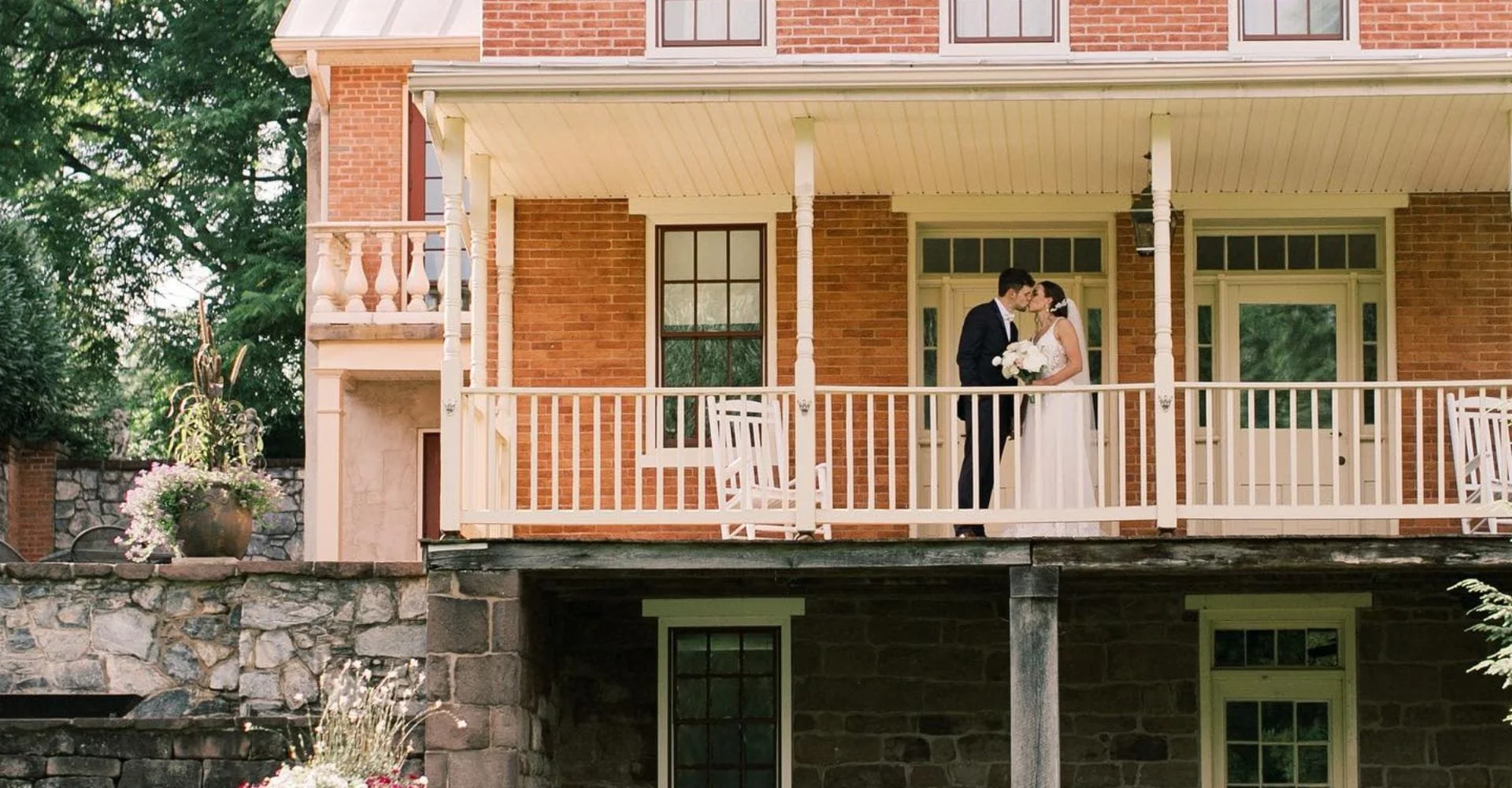 A couple in wedding attire embraces on a charming brick house porch. The scene is serene, with lush greenery and rustic stonework enhancing the romantic atmosphere.
