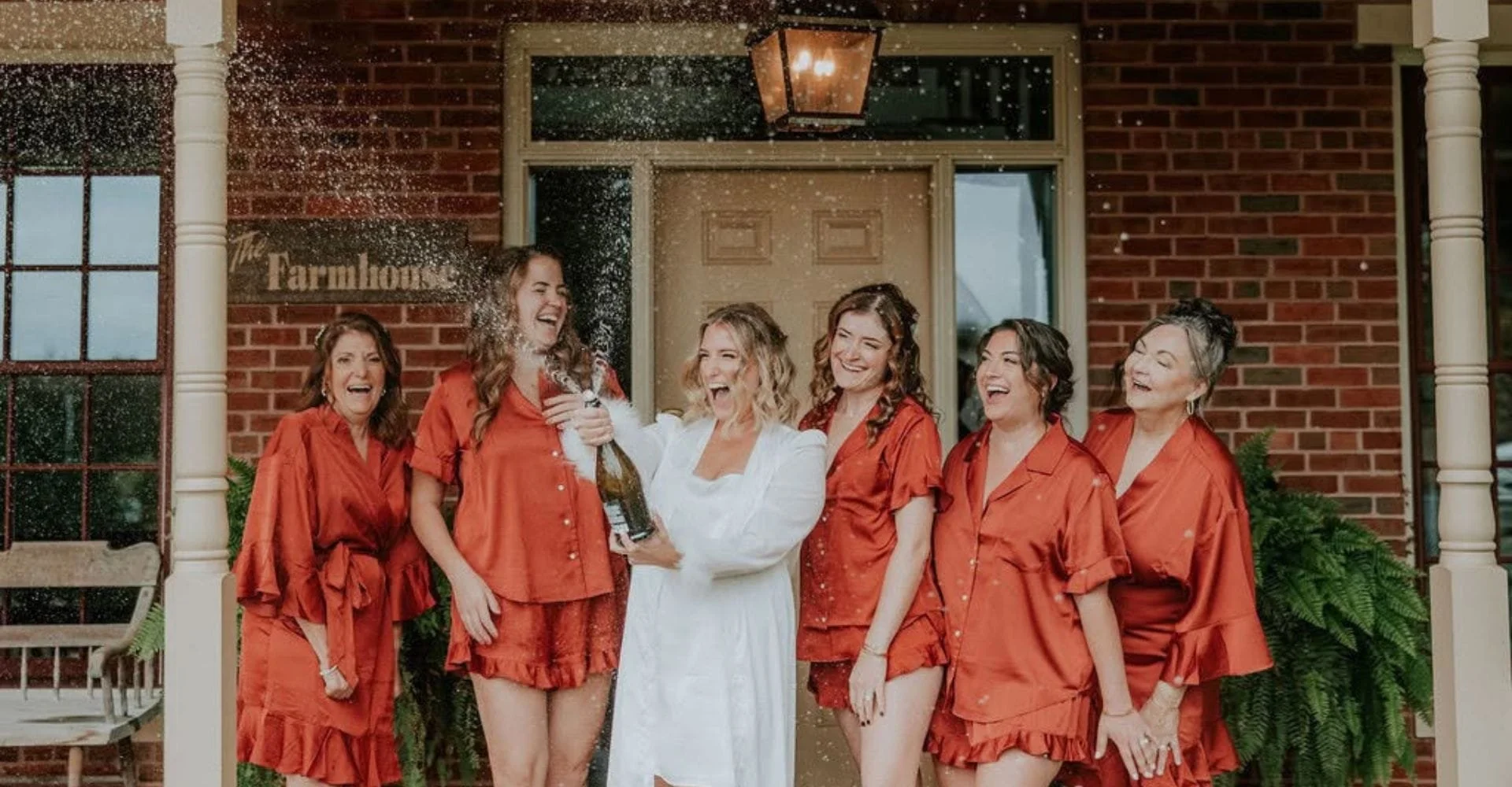 Bridesmaids in red robes pose together outside a charming brick building, smiling and enjoying the moment.