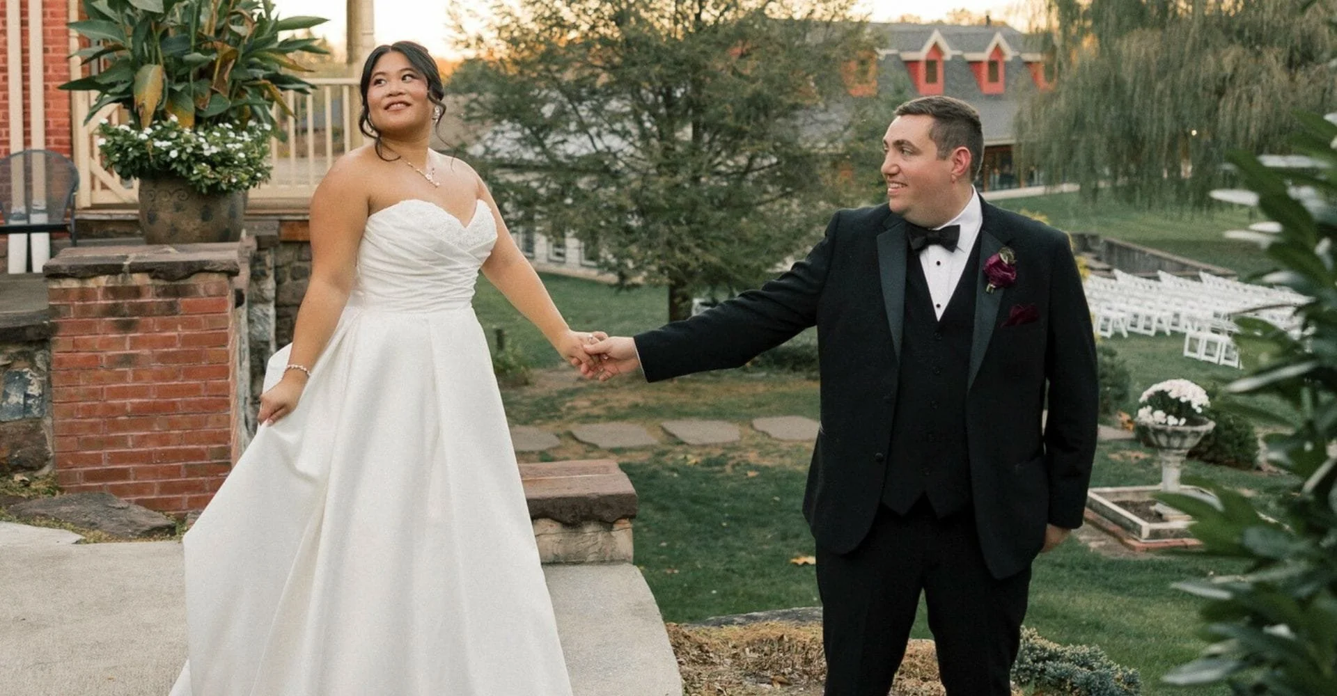 Bride in white gown and groom in black tuxedo holding hands outdoors. Bride smiles upward, groom looks at her. Romantic and joyful atmosphere.