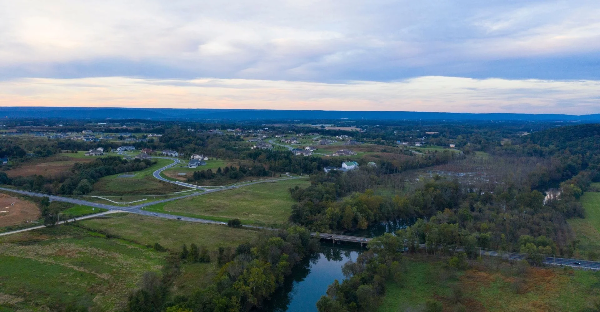 Aerial view of a vast, serene landscape at dusk, featuring winding roads through green fields, scattered houses, dense trees, and a calm river reflecting the sky.