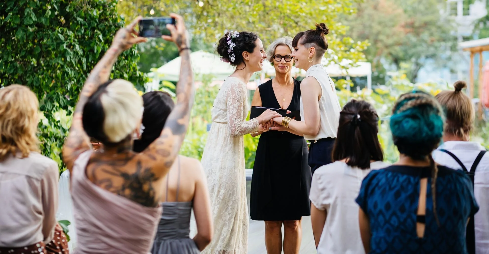 A wedding ceremony outdoors with two brides, one in a lace dress and the other in a blouse and skirt, holding hands, smiling, surrounded by guests.