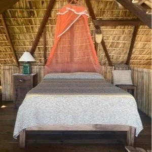 A rustic bedroom with a bed, orange mosquito net, and wooden furniture inside an A-frame structure.