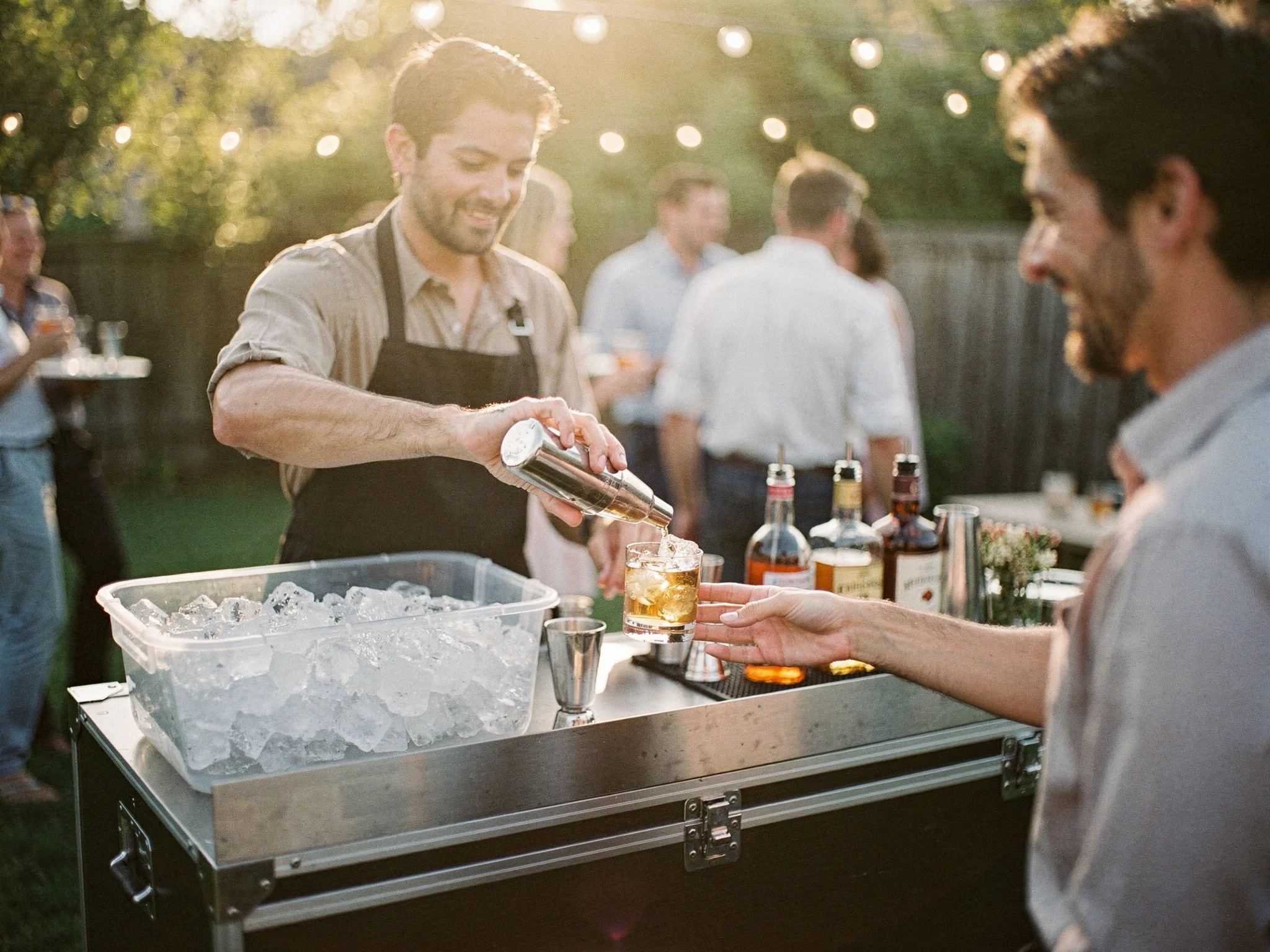 A bartender in a black apron pours a cocktail at an outdoor party bar with a large ice bin on the counter