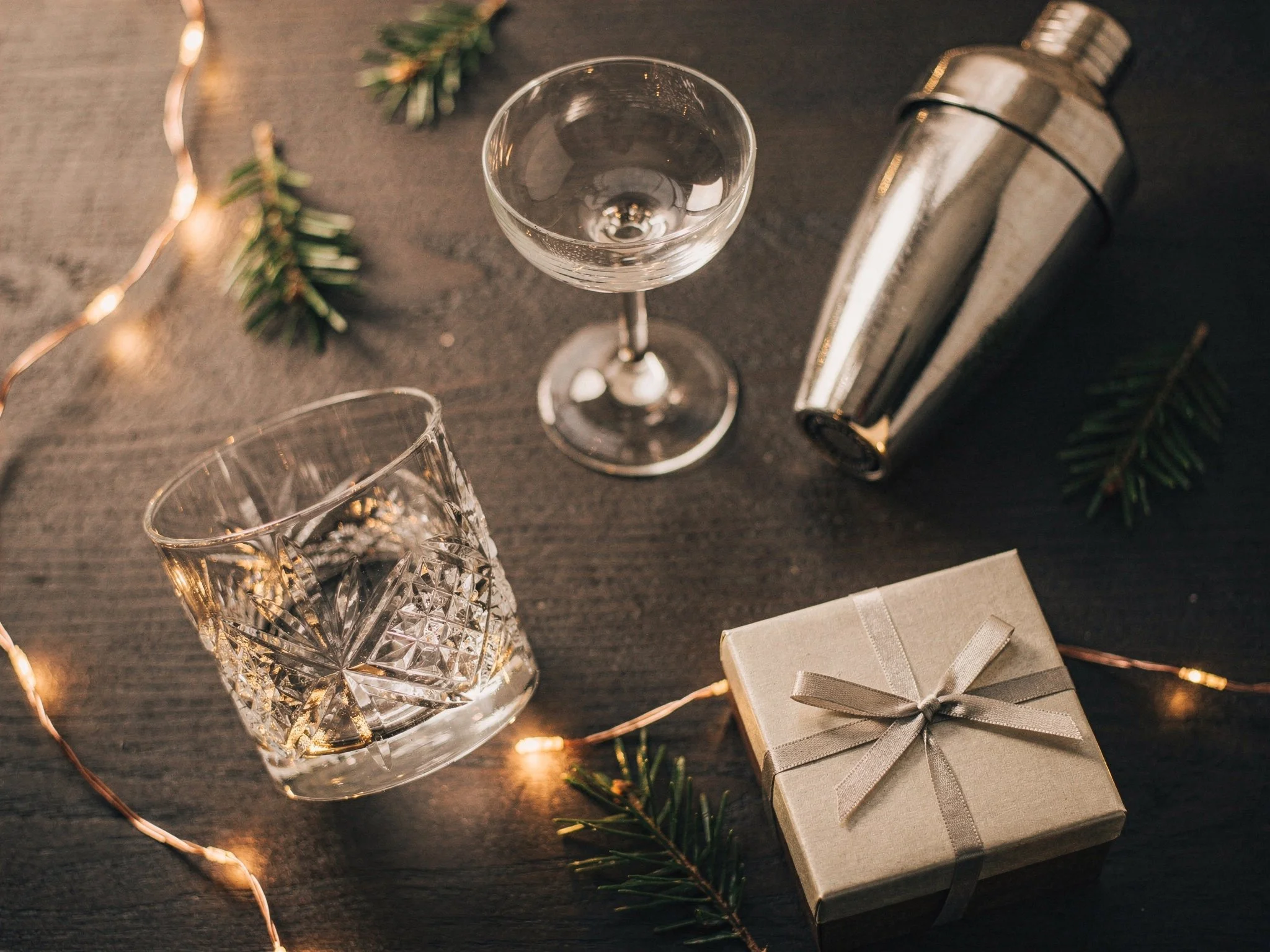 A rocks glass, a coupe glass, a cocktail shaker, and a wrapped gift box styled for a holiday barware gift guide