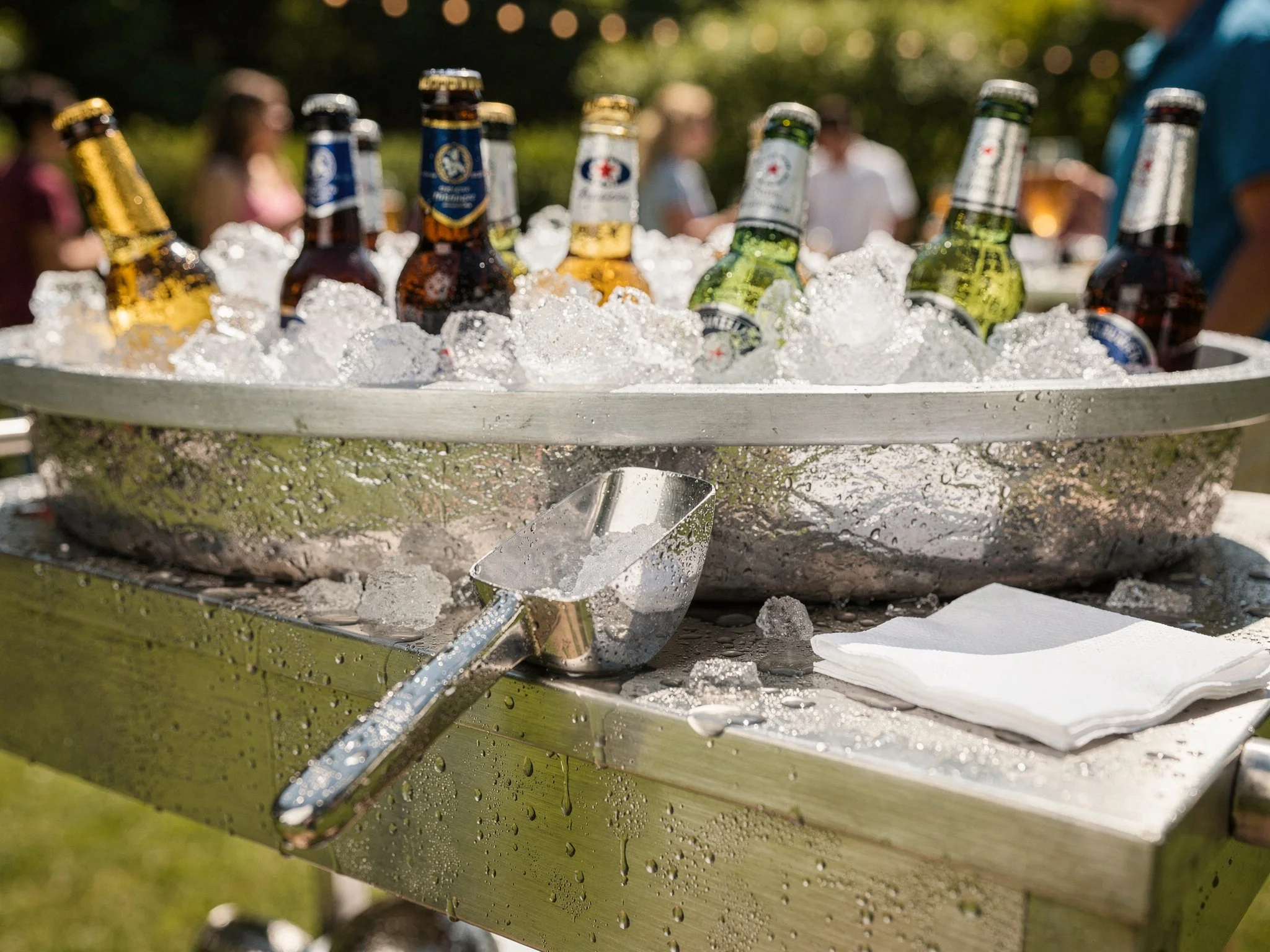 A stainless steel outdoor bar cart packed with crushed ice and chilled bottles, condensation beading in bright sunlight