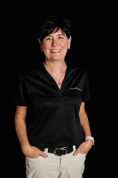 A woman with short dark hair smiling, wearing a black polo shirt with a logo, white pants, and a watch, standing against a black background.